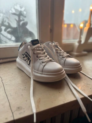 Vita sneakers från Karl Lagerfeld - Säljer ett par vita sneakers från Karl Lagerfeld med svart detalj på hälen och coolt kattmärke på sidan. Skorna har snörning, platt sula och rund tå. Tillverkade i skinn med en chunky sula som ger en modern vibe. Perfekta för dig som gillar stilrena men ändå lekfulla detaljer.