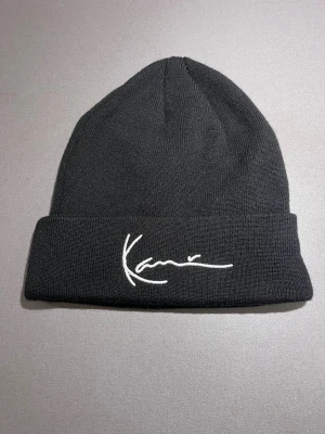Svart mössa från Karl Kani - Svart mössa från Karl Kani med broderad vit logga framtill. Klassisk form med uppvikt kant och mjukt stickat material som sitter skönt på huvudet. One size, men i andra märken har jag M/L. Helt ny, aldrig använd! Säljs på flera sidor, först till kvarn. 