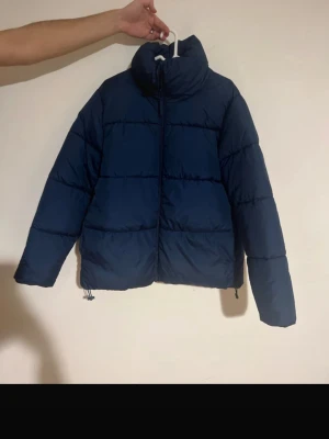  pufferjacka från Cubus - Säljer en mörkblå pufferjacka från Cubus i storlek M. Jackan har hög krage, dragkedja framtill och justerbar nederkant med snörning. Perfekt för kalla dagar och har en clean, stilren look som funkar till många outfits.