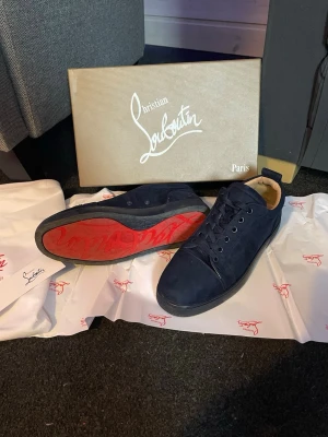 Mörkblå Christian Louboutin sneakers - Snygga mörkblå sneakers från Christian Louboutin med klassisk röd sula och diskret logga på sidan. Skorna har rund tå, snörning och är tillverkade i mocka med beige insida. Perfekt för dig som gillar exklusiva och stilrena detaljer.