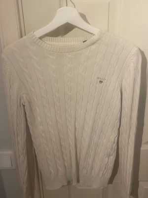 Vit kabelstickad tröja från GANT - Säljer en vit kabelstickad tröja från GANT med broderad logga på bröstet. Tröjan har rund hals, ribbade muddar och lång ärm. Perfekt för dig som gillar klassisk och stilren design med lite preppy känsla.