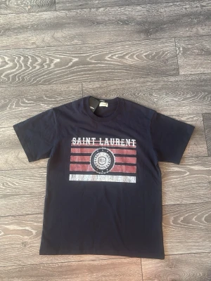 Mörkblå Saint Laurent t-shirt  - Snygg mörkblå t-shirt från Saint Laurent med coolt tryck framtill i vitt och rött. Klassisk rund hals och korta ärmar. Tillverkad i mjuk bomull som känns skön mot huden. Perfekt för dig som vill ha en statement tee med exklusiv känsla.
