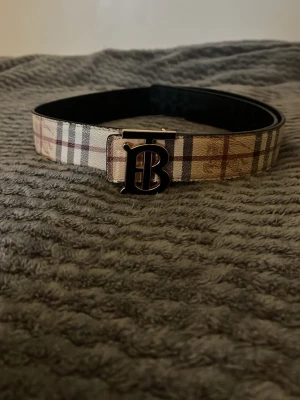 Rutig Burberry-väst med B-spänne - Säljer en snygg väst från Burberry med klassiskt rutig design i beige, brun och svart. Västen har ett stilrent B-spänne i metall framtill och är tillverkad i skinn. Insidan är svart och utsidan har det ikoniska rutmönstret. Perfekt för dig som vill ha en statement piece.