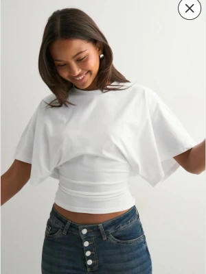 Vit croppad topp med vida ärmar - Säljer en vit croppad topp med breda, korta ärmar och rund halsringning. Toppen har en figurnära passform över midjan och är tillverkad i ett mjukt bomullsmaterial. Perfekt för en trendig och avslappnad look.