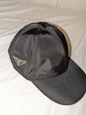 Svart keps från Prada i nylon - Säljer en stilren svart keps från Prada med den ikoniska metall-loggan på sidan. Kepsen är tillverkad i glansigt nylonmaterial och har en klassisk böjd skärm. Perfekt för dig som gillar exklusiva accessoarer och vill sticka ut med designerplagg.