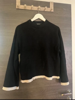 Svart stickad tröja från Gina Tricot - Svart stickad tröja från Gina Tricot med ribbade beige detaljer vid ärmslut och nederkant. Tröjan har lång ärm, rund hals och en loose passform. Perfekt för dig som gillar en chill och enkel stil med lite extra detalj på kanterna.