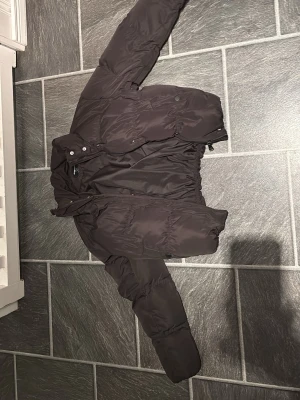 Svart pufferjacka från FB Sister - Svart pufferjacka från FB Sister i storlek S. Jackan har en hög krage, dragkedja och tryckknappar framtill. Den är vadderad och har långa ärmar med elastiska detaljer vid handlederna. Perfekt för kalla dagar och har en clean, enkel look.