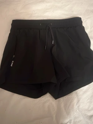 Svarta träningsshorts från ICANIWILL - Säljer ett par svarta träningsshorts från ICANIWILL med elastisk midja och snörning. Shortsen har en loose passform, innerbyxa i mesh och praktisk dragkedja på sidan. Perfekta för gymmet eller löpning, riktigt sköna och stilrena.
