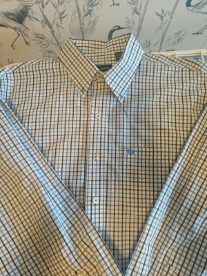 Rutig skjorta från IZOD i blått och vitt - Snygg skjorta från IZOD med klassiskt rutigt mönster i blått och vitt. Skjortan har knappar framtill, krage och en liten broderad logga på bröstfickan. Perfekt för dig som gillar stilrena och tidlösa plagg. Materialet känns lätt och bekvämt.