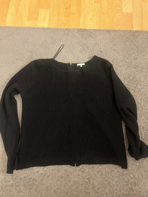 Svart stickad tröja med dragkedja - Säljer en svart stickad tröja med lång ärm och snygg guldfärgad dragkedja längs ryggen. Tröjan har en enkel, rak passform och är perfekt för kyligare dagar. Stilren och lätt att matcha med olika outfits.