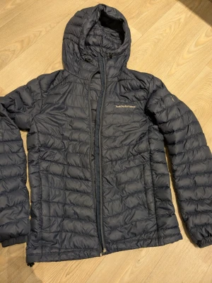 Mörkblå Peak Performance dunjacka S - Peak Performance Frost DH dunjacka i mörkblått med huva och dragkedja. Jackan är quiltad och tillverkad i lätt syntetmaterial med Pertex Quantum-tyg. Perfekt för kalla dagar och har en clean, sportig look. Saknar löpare på dragkedjan, ny kostar ca 60kr. Två små lagningar (se bild) Nypris 3199kr