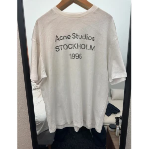 Vit Acne Studios T-shirt  - Vit t-shirt från Acne Studios med svart tryck på ryggen där det står 'Acne Studios STOCKHOLM 1996'. Modellen är klassisk med rund hals och korta ärmar, och har en loose passform. Materialet är mjuk bomull som känns skönt mot huden. Stor i storleken så den passar även M och L