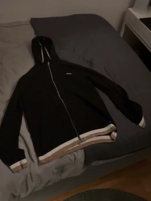 Svart hoodie från BOSS med dragkedja, Läs beskrivningen! - Svart hoodie från BOSS i storlek XS skulle jag säga med hel dragkedja framtill och huva. Tröjan har vita och beige ränder vid muddar och nederkant, samt BOSS-logga på bröstet. Perfekt för chill dagar och snygg streetstil. Denna kofta säljs inte längre så passa på innan den säljs!!. Köptes för 1400 säls för 500🫡.