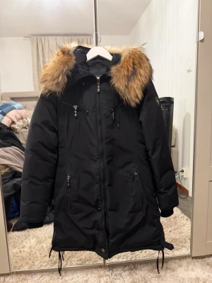 Svart parkajacka med päls från Hollies - Säljer en svart parkajacka från Hollies med fluffig päls runt huvan. Jackan har flera dragkedjor framtill, långa ärmar och snörning vid nederkanten. Perfekt för kalla dagar och har en snygg, klassisk look. Storlek 38, men modellen är tight i passformen – passar även dig som vanligtvis har storlek 36.