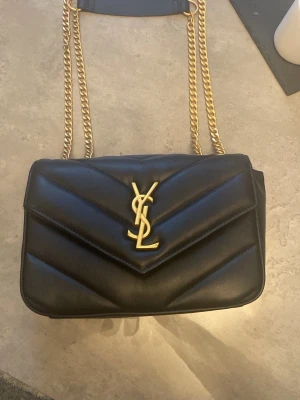 Svart Saint Laurent axelväska i skinn - Säljer en svart axelväska från Saint Laurent med quiltad design och guldiga kedjor. Väskan har YSL-logga i guld på framsidan och flera fack inuti. Medföljer matchande svart plånbok med knappstängning. Perfekt för dig som gillar lyxiga detaljer och stilren look. Har aldrig använt eftersom att jag fick den i present och jag har inte den stilen!!!!