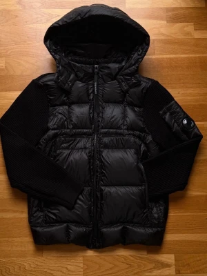 Svart cardigan från C.P. Company - Säljer en svart pufferjacka från C.P. Company med stickade ärmar och huva. Jackan har dragkedja framtill, ribbade muddar och en ikonisk linsdetalj på ärmen. Materialmix av polyester och stickad ull ger både värme och cool stil.
