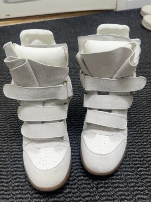 Isabel Marant vita sneakers med kardborre - Säljer ett par vita Isabel Marant sneakers med högt skaft och flera breda kardborreband. Skorna har en rund tå, platt sula i gummi och är tillverkade i mocka och skinn. De har en chunky design och coola detaljer som ger en unik look.