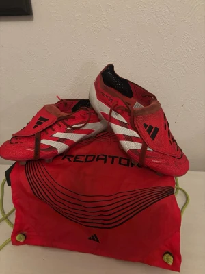 Adidas Predator röda fotbollsskor - Säljer ett par Adidas Predator fotbollsskor i rött med vita och svarta detaljer. Skorna har snörning, platt sula med dobbar och är tillverkade i syntetmaterial. Medföljer en matchande röd Predator-påse. Perfekt för dig som vill sticka ut på planen. Samt låda till skorna. Skorna är inte använda så mycket 8/10. Köpta för 3000 kr och är i storlek 42 2/3
