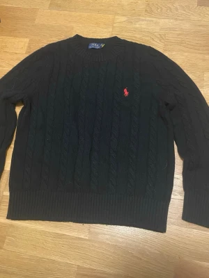 Svart kabelstickad tröja Polo Ralph Lauren - Svart kabelstickad tröja från Polo Ralph Lauren med röd broderad logga på bröstet. Tröjan har rund hals, ribbade muddar och är långärmad. Perfekt för dig som gillar klassisk stil med en modern touch.