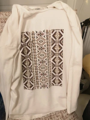 Vit långärmad tröja LC Waikiki XXL - Vit långärmad tröja från LC Waikiki i storlek XXL. Framsidan har ett stort geometriskt mönster i brunt, beige och guld med detaljer som ger en cool vibe. Mjuk och skön bomullskänsla, perfekt för dig som gillar stilrena men ändå unika plagg.