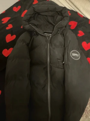 Svart pufferjacka Supply & Demand - Säljer en svart pufferjacka från Supply & Demand med huva och dragkedja framtill. Jackan har en patch med logga på ärmen och en clean, oversized look. Perfekt för kalla dagar när du vill hålla stilen och värmen.