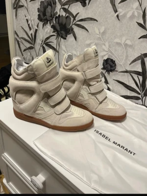 Isabel Marant vita höga sneakers - Säljer ett par vita höga sneakers från Isabel Marant med beige mockadetaljer och bruna gummisulor. Skorna har flera kardborreband, rund tå och inbyggd kilklack för extra höjd. Perfekt för dig som gillar statement-skor med cool design.