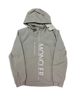 Grå vindjacka från Moncler med huva - Snygg grå vindjacka från Moncler med stor logga längs dragkedjan och praktisk huva. Jackan har elastiska muddar, bröstficka med dragkedja och Moncler-märke på ärmen. Perfekt för dig som gillar stilren design och funktion.