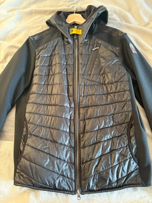 Svart quiltad jacka från Parajumpers - Svart jacka från Parajumpers med quiltad front och huva. Snygga detaljer som gul rem vid halsen och patch på ärmen. Jackan har dragkedja, bröstficka med zip och sidofickor. Materialmix med polyester och syntet, perfekt för kyliga dagar. Jackan är köpt för 4 månader sedan. Den är helt ny utan prislapp, inga skador. Köpt för 4499 kr på Johnells (bild på kvitto kan visas i meddelande). 