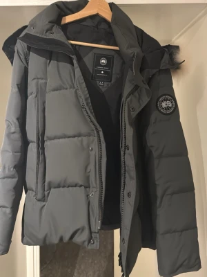 Mörkgrå Canada Goose pufferjacka - Säljer en mörkgrå pufferjacka från Canada Goose med ikonisk patch på ärmen och stor huva med äkta päls.Går även att ta bort! Jackan har dragkedja och knappar framtill, quiltad design och är långärmad. Perfekt för kalla vinterdagar och riktigt snygg streetstil. Bara höra av sig vid frågor😊