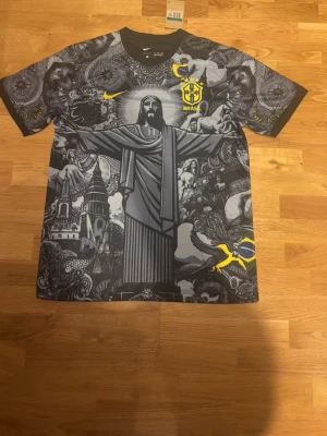 Nike Brasil t-shirt med grafiskt tryck - Säljer en svart och grå Nike t-shirt med Brasil-logga och stort grafiskt tryck av Kristusstatyn och brasilianska motiv. Gula detaljer och Nike Swoosh på bröstet. Materialet är mjukt och lätt, perfekt för dig som gillar unika och sportiga plagg.