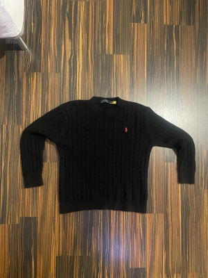 Svart stickad tröja Polo Ralph Lauren - Svart kabelstickad tröja från Polo Ralph Lauren i storlek M. Klassisk rund hals och långärmad modell med röd broderad logga på bröstet. Tröjan är gjord i mjukt material och har ribbade muddar vid ärmslut och nederkant. Perfekt för kyliga dagar.