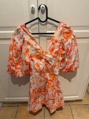 Blommig kortklänning med puffärm XS - Superfin kortklänning med blommigt mönster i orange, rosa och vita toner. Klänningen har puffiga kortärmar, v-ringad hals och en stor rosett i ryggen. 