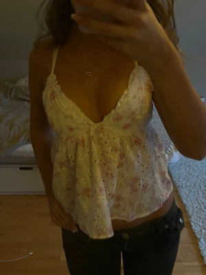 Vit blommig linnetopp med broderi - Säljer en söt vit linnetopp med rosa blommigt mönster och broderade hålblommor. Toppen har smala axelband som korsas i ryggen och en v-ringad front. Materialet är lätt och luftigt, perfekt för varma dagar. Toppen är ärmlös.