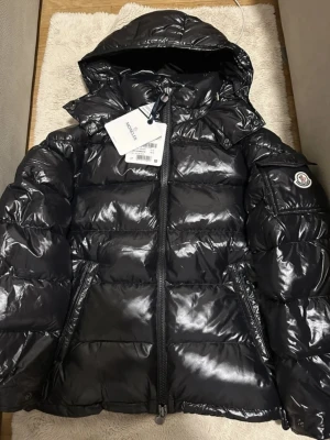 Moncler maya ev byte - Säljer en svart, glansig pufferjacka från Moncler med huva och dragkedja framtill. Jackan har en ficka med Moncler-logga på ärmen och quiltad design. Perfekt för kalla dagar och har en riktigt snygg streetstil. Ev byte