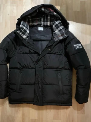 Svart pufferjacka från Burberry - Svart pufferjacka från Burberry med huva och rutigt foder i svart, vitt och rött. Jackan har dragkedja och knappar framtill samt två fickor med dragkedja. Märke med 'Burberry London England' på ärmen. Perfekt för kalla dagar.