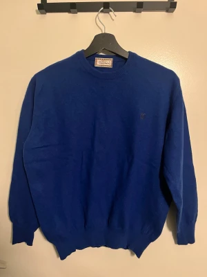 Blå stickad tröja från Thomas Burberry - Snygg blå stickad tröja från Thomas Burberry med rund hals och diskret broderad logga på bröstet. Tröjan har ribbade muddar vid ärmslut och nederkant, vilket ger en klassisk och stilren look. Perfekt för dig som gillar enkel och tidlös design. Aldrig använd och ny skick