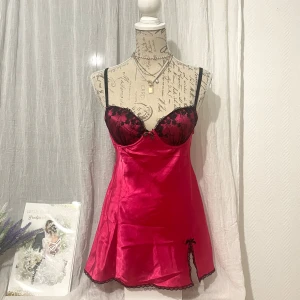Rosa satin babydoll med spetsdetaljer - Supersnygg kort babydoll-klänning i stark rosa satin med svarta spetsdetaljer på kuporna, liten rosett framtill och slits med rosett på sidan. Storlek S, passar 34-36, byst 83-88cm. Dockan är i storlek 36, byst 86cm.