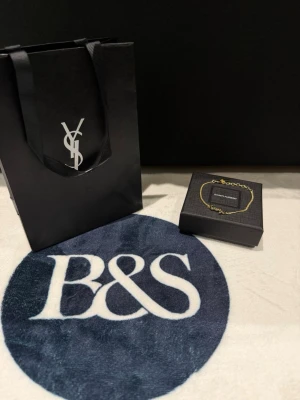 YSL Yves Saint Laurent gold bracelet -Unisex - Exklusivt armband från Saint Laurent i guldfärgad metall med den ikoniska YSL-loggan framtill. Kedjan är tunn och elegant med justerbar längd. Kommer i original ask och presentpåse med Saint Laurent-tryck. Perfekt accessoar för dig som gillar lyxiga detaljer.