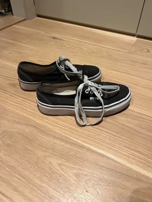 Svarta Vans sneakers - Klassiska svarta Vans sneakers med vit sula. Skorna har rund tå, lågt skaft och tydliga vita sömmar som kontrast. Ikonisk Vans-logga baktill och på sidan. Perfekta för en avslappnad streetstil.