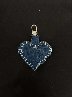 Hjärtfomad nyckelring med vit söm - Unik nyckelring i hjärtform, sydd i mörkblått jeanstyg med grova vita sömmar längs kanterna. Nyckelringen har en metallhake upptill för enkel fastsättning. Perfekt för dig som gillar kreativ och personlig stil.