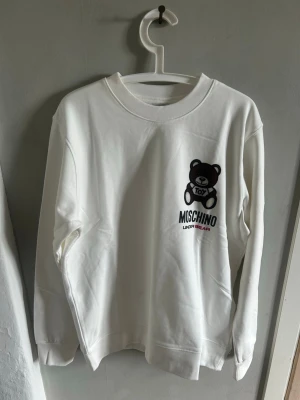 Vit Moschino sweatshirt  - Säljer en vit sweatshirt från Moschino med ett stort tryck av den ikoniska björnen och texten 'Toy' på bröstet. Tröjan har rund hals, långa ärmar och är gjord i mjuk bomull. Perfekt för dig som gillar streetstyle och coola designerplagg.