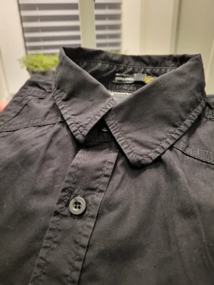 Svart skjorta från Jack & Jones - Svart skjorta från Jack & Jones med klassisk krage, två bröstfickor med knappar och snygg broderad logga på ena sidan. Skjortan har långa ärmar och stängs med knappar framtill. Insidan av kragen har ett rutigt svartvitt mönster. Perfekt till en clean och stilren look.