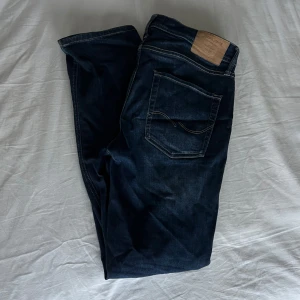Jack and Jones - Säljer ett par Jack&Jones jeans i mörkblå tvätt. Klassisk raka ben och normal/lite slim passform. Jeansen har snygga kontrastsömmar och ett brunt läderpatch bak i midjan.