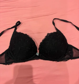 Svart spets-bh med rosett - Snygg svart bh med spetsdetaljer och en liten rosett med silverfärgad fjäril mellan kuporna. Kuporna är vadderade och har meshpartier på sidorna. Perfekt för dig som gillar stilrena och feminina underkläder.