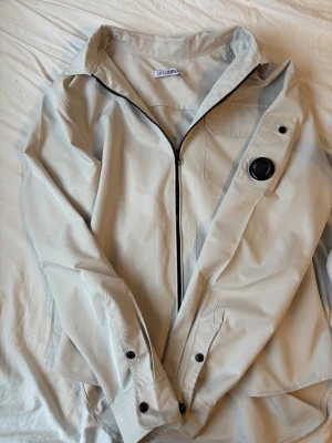 Beige overshirt från C.P. Company - Säljer en clean beige overshirt från C.P. Company med ikonisk linsdetalj på ärmen. Jackan har dragkedja framtill, bröstficka, knappdetaljer vid ärmslut. Perfekt för dig som gillar streetwear och vill ha något stilrent. Vill få den sålt så snabbt som möjligt kan därav gå ner något i pris nästan perfekt skick då den knappt kommit till användning