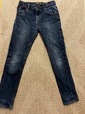 Mörkblå jeans från Lyle & Scott Junior - Snygga mörkblå jeans från Lyle & Scott Junior med klassisk femficksdesign och gul kontrastsöm. Jeansen har rak passform och är tillverkade i slitstarkt denim med lätt tvättad look. Perfekt för dig som gillar stilrena och tidlösa jeans.