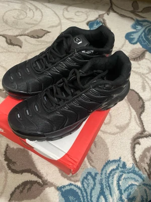Nike Air Max Plus svarta sneakers - Nike Air Max Plus sneakers i helsvart färg med mesh och syntetpaneler, ikoniska vågiga detaljer och synlig Air Max-dämpning i sulan. Rund tå, snörning och platt sula. Snygg och sportig look som passar perfekt till streetwear.