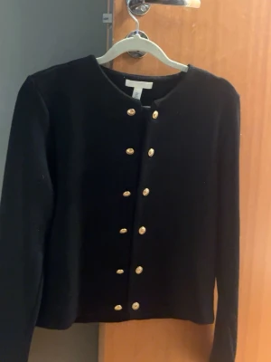 Svart kofta med guldknappar från H&M - Svart kofta från H&M med rund hals och snygga guldknappar framtill. Långärmad och i mjukt material som känns skönt mot huden. Använd cirka 1-2 gånger precis som ny