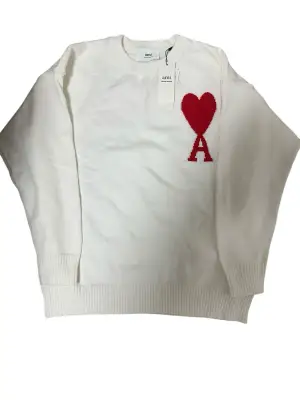 Vit stickad tröja från Ami med rött hjärta och bokstaven A broderat på bröstet. Tröjan har rund hals, ribbade muddar och är långärmad. Perfekt för dig som gillar stilrena detaljer och vill ha något som sticker ut lite extra.