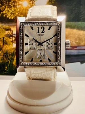 Chick klocka från Guess - Säljer en stilren fyrkantig klocka från Guess med vit, krokopräglad rem och glittrande stenar runt boetten. Urtavlan är silverfärgad med flera små ur och tydliga svarta markeringar. Klockan har en lyxig vibe och passar perfekt för dig som gillar att sticka ut.